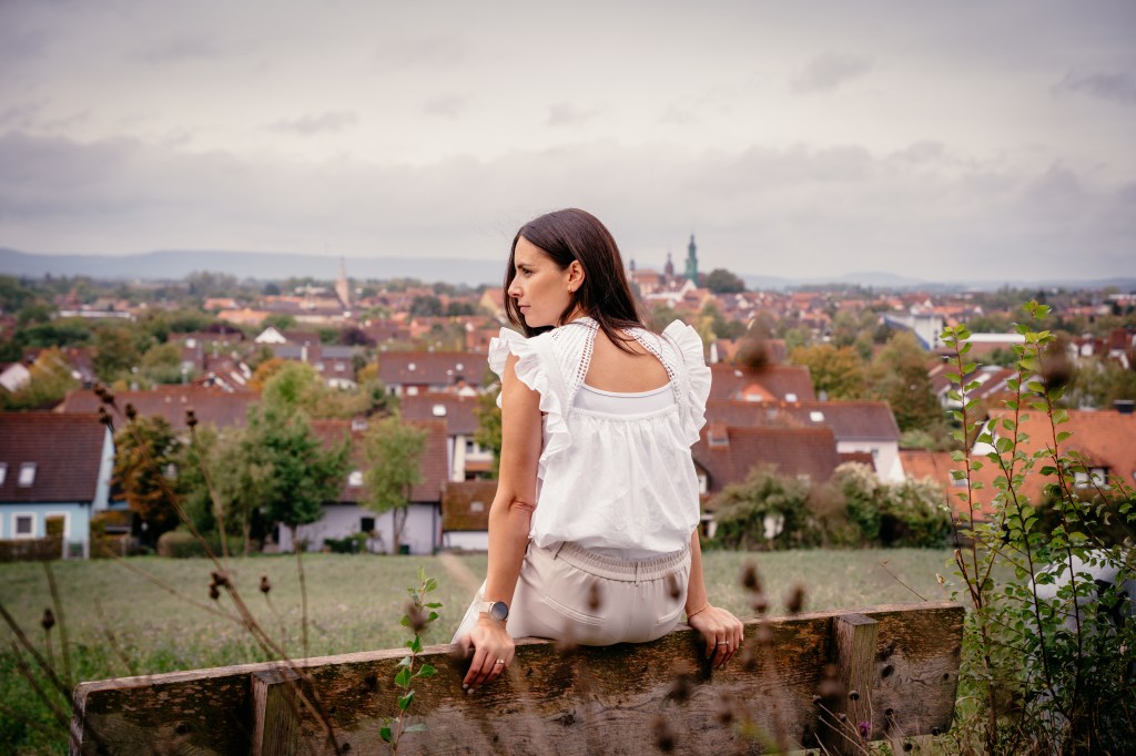 Eine Frau mit langen, braunen Haaren sitzt auf einer Bank und blickt nachdenklich auf eine Stadtlandschaft mit roten Dächern im Hintergrund. Der Himmel ist bewölkt, und die Umgebung wirkt grün und ländlich.
